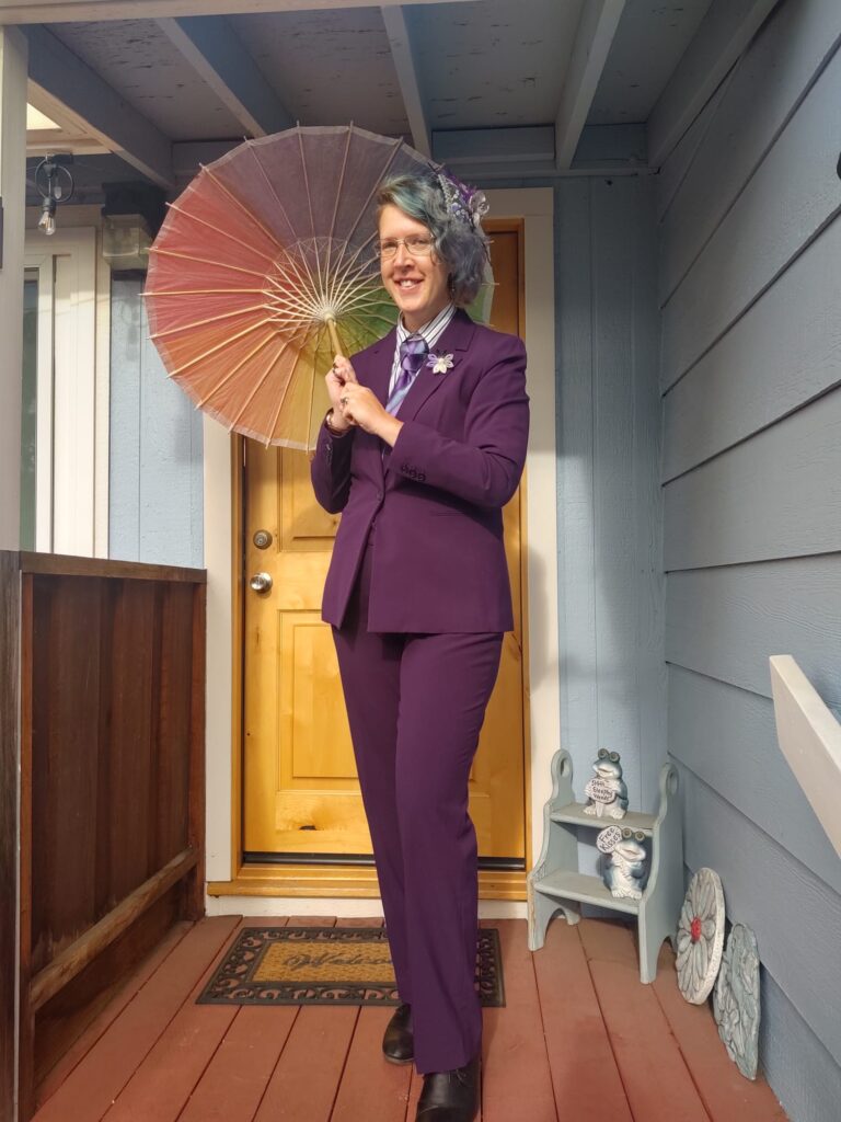 Een persoon met blauw haar en een bril staat in een paars pak voor een voordeur. Ze houdt een parasol in regenboogkleuren over zir rechter schouder en kijkt lachend de camera in.