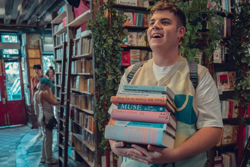 Het personages Isaac staat in een boekenwinkel. Hij houdt een stapel boeken vast en kijkt blij weg van de camera.